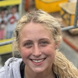 Eine junge blonde Frau in grauer Kapuzenjacke lächelt fröhlich, während sie auf einem Dach mit roten Ziegeln arbeitet. Im Hintergrund sind Baugerüste und Baustellen-Equipment.