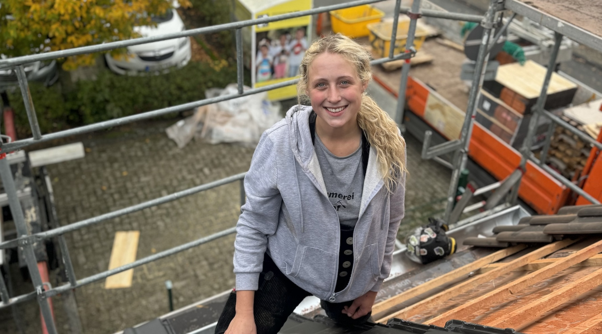 Eine junge blonde Frau in grauer Kapuzenjacke lächelt fröhlich, während sie auf einem Dach mit roten Ziegeln arbeitet. Im Hintergrund sind Baugerüste und Baustellen-Equipment.
