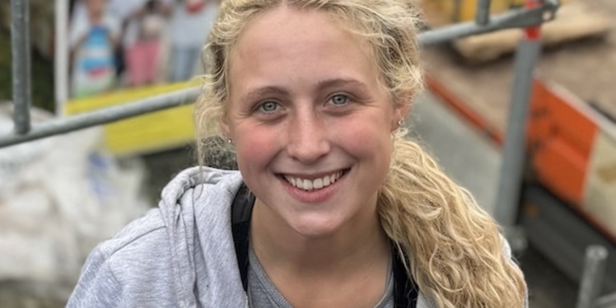 Eine junge blonde Frau in grauer Kapuzenjacke lächelt fröhlich, während sie auf einem Dach mit roten Ziegeln arbeitet. Im Hintergrund sind Baugerüste und Baustellen-Equipment.