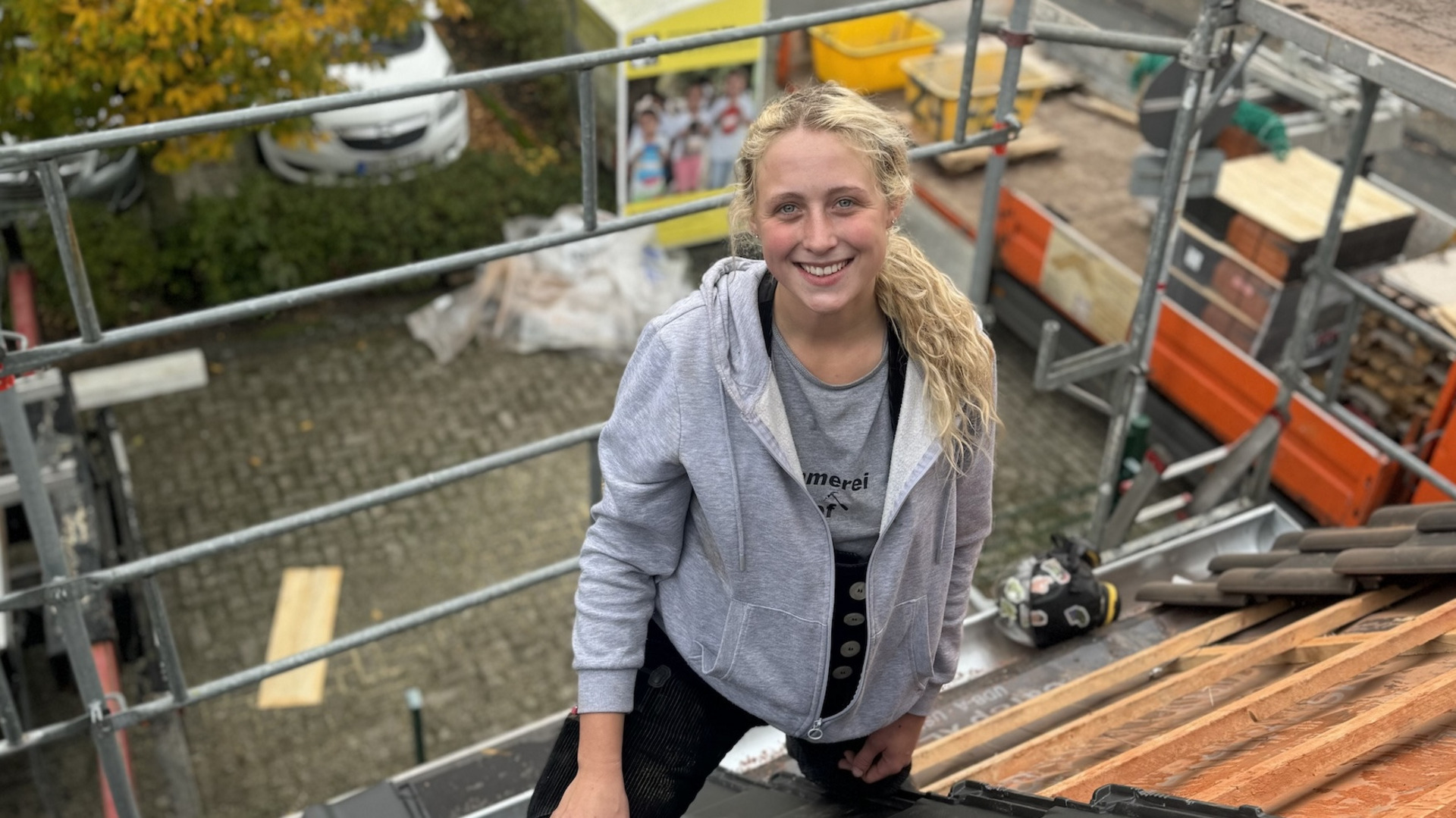 Eine junge blonde Frau in grauer Kapuzenjacke lächelt fröhlich, während sie auf einem Dach mit roten Ziegeln arbeitet. Im Hintergrund sind Baugerüste und Baustellen-Equipment.