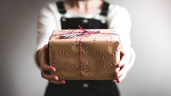 Eine festlich gekleidete Frau hält ein schön verpacktes Geschenk in die Kamera.