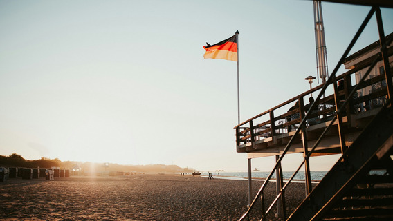 Das Bild zeigt einen Sandstrand bei Sonnenuntergang, auf dem im Vordergrund ein Gebäude auf Stelzen mit einer Treppe zu sehen ist. Eine deutsche Flagge weht auf einem hohen Fahnenmast neben dem Gebäude. Im Hintergrund sind Strandkörbe entlang des Strandes aufgereiht, und einige Menschen spazieren entlang der Wasserkante. Die Sonne steht tief am Himmel und taucht die Szene in ein warmes, goldenes Licht. Das Bild vermittelt eine ruhige und entspannte Atmosphäre.