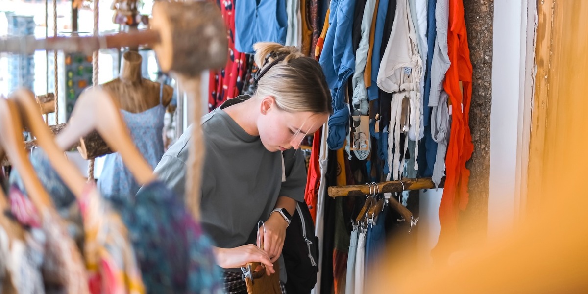 Eine Person stöbert in einem Secondhand-Laden durch einen Kleiderständer mit bunter Kleidung und betrachtet ein Kleidungsstück genauer.