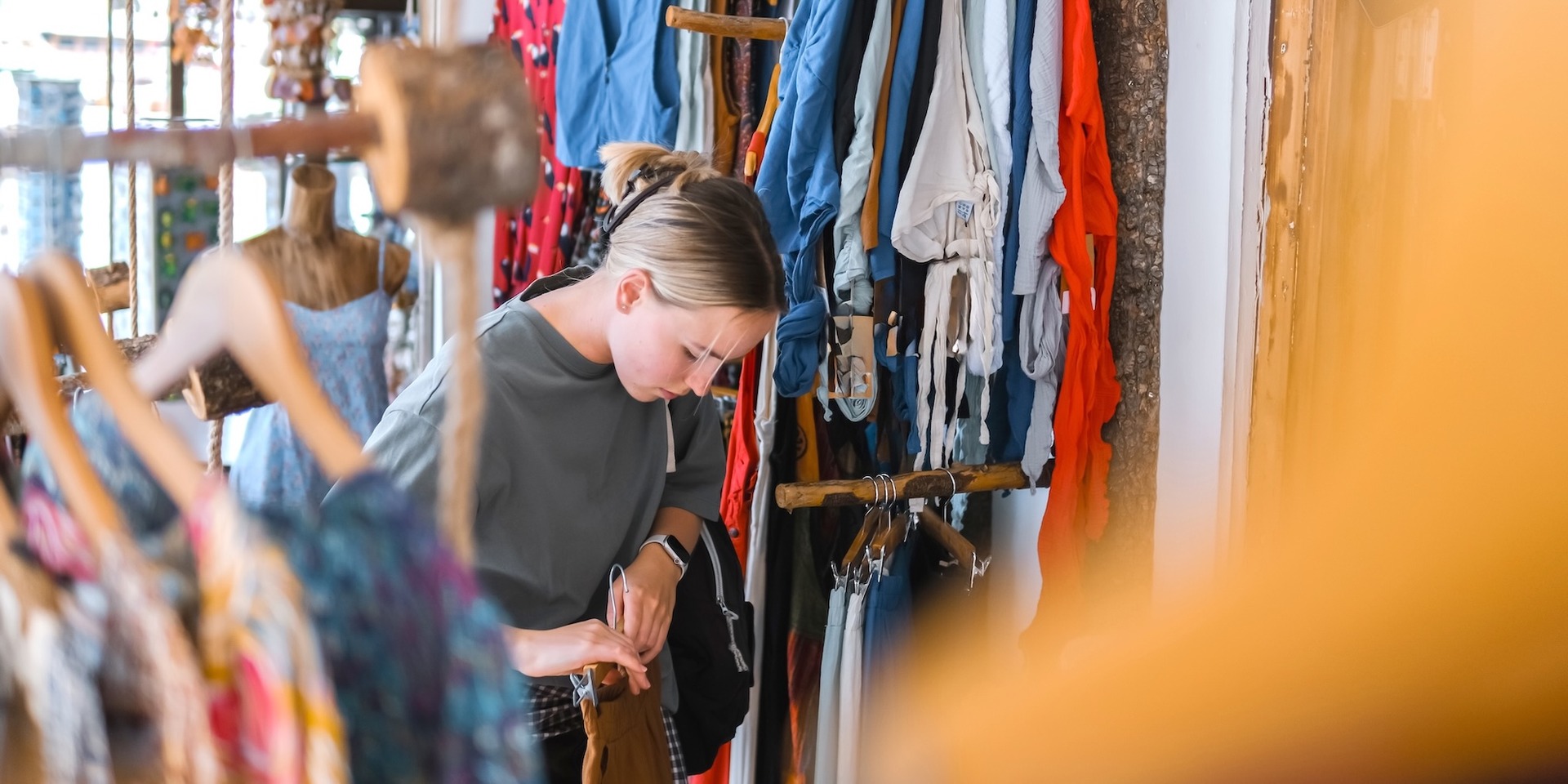 Eine Person stöbert in einem Secondhand-Laden durch einen Kleiderständer mit bunter Kleidung und betrachtet ein Kleidungsstück genauer.