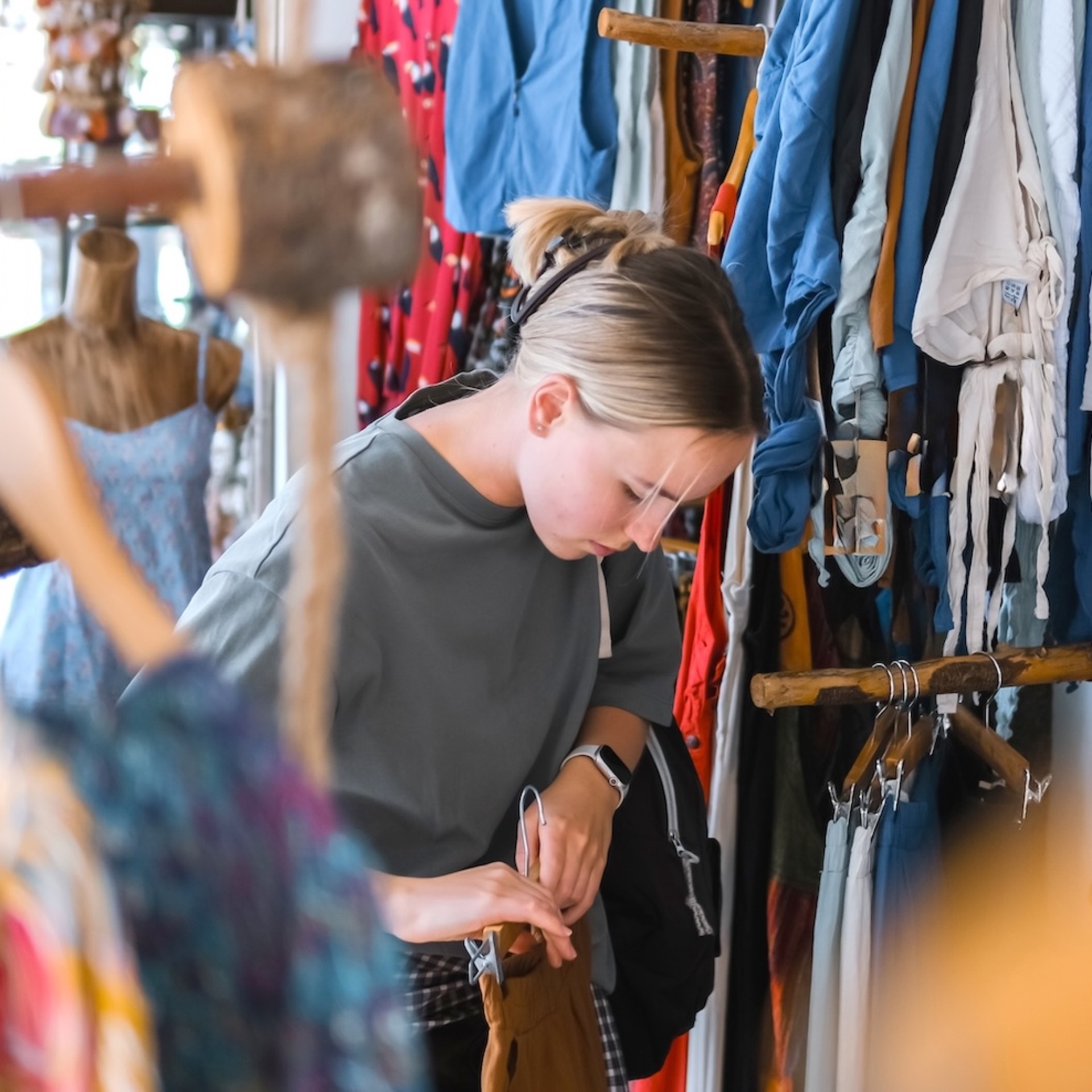 Eine Person stöbert in einem Secondhand-Laden durch einen Kleiderständer mit bunter Kleidung und betrachtet ein Kleidungsstück genauer.