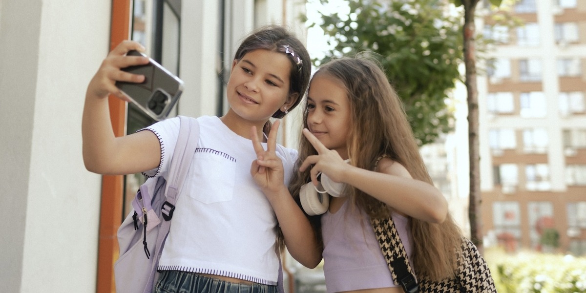 wei junge Mädchen mit Schulrucksäcken machen draußen vor einem Wohngebäude ein Selfie mit einem Smartphone und zeigen dabei Peace-Zeichen. Im Hintergrund sind Bäume und Wohnhäuser zu sehen.