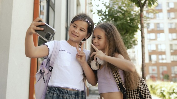 wei junge Mädchen mit Schulrucksäcken machen draußen vor einem Wohngebäude ein Selfie mit einem Smartphone und zeigen dabei Peace-Zeichen. Im Hintergrund sind Bäume und Wohnhäuser zu sehen.