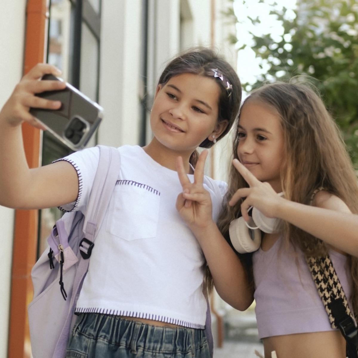 wei junge Mädchen mit Schulrucksäcken machen draußen vor einem Wohngebäude ein Selfie mit einem Smartphone und zeigen dabei Peace-Zeichen. Im Hintergrund sind Bäume und Wohnhäuser zu sehen.