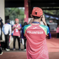 Eine Person mit der Aufschrift "Volunteer" (engl. "Freiwillige:r") steht mit dem Rücken zu uns und schießt ein Foto einer Szene im Hintergrund