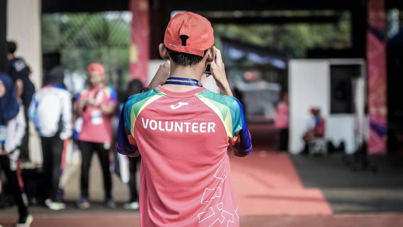 Eine Person mit der Aufschrift "Volunteer" (engl. "Freiwillige:r") steht mit dem Rücken zu uns und schießt ein Foto einer Szene im Hintergrund