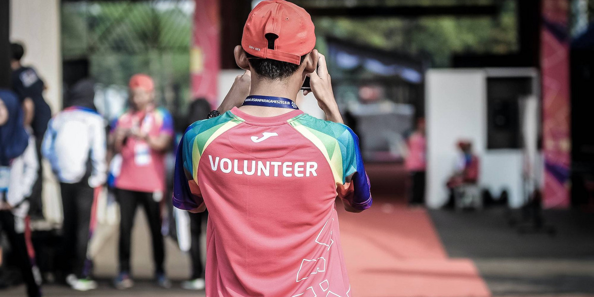 Eine Person mit der Aufschrift "Volunteer" (engl. "Freiwillige:r") steht mit dem Rücken zu uns und schießt ein Foto einer Szene im Hintergrund
