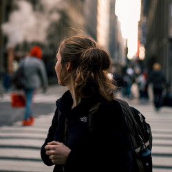 Eine junge Person steht im Vordergrund des Bildes, den Blick abgewendet Richtung Stadt. Im Hintergrund sind in der Unschärfe eine Kreuzung mit weiteren Menschen und Leuchtreklame zu erkennen.
