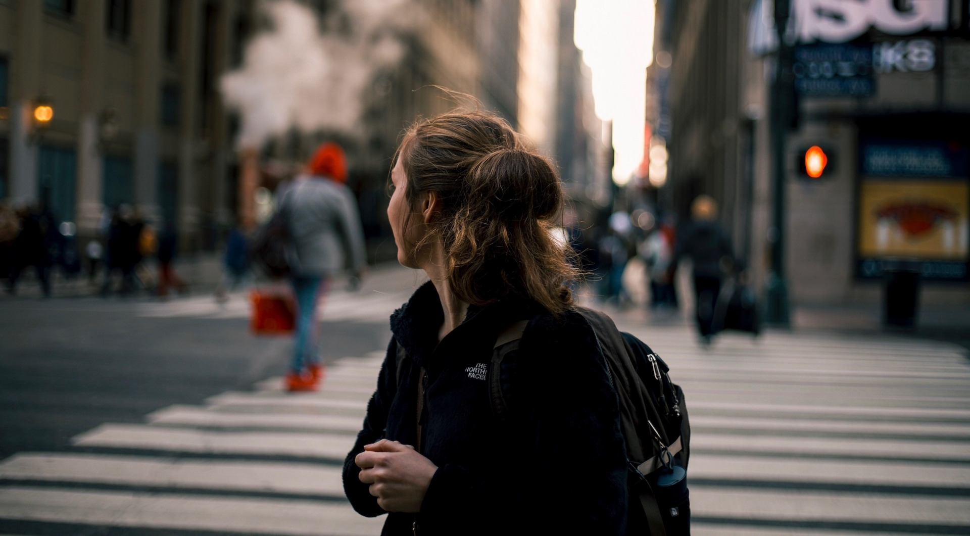 Eine junge Person steht im Vordergrund des Bildes, den Blick abgewendet Richtung Stadt. Im Hintergrund sind in der Unschärfe eine Kreuzung mit weiteren Menschen und Leuchtreklame zu erkennen.