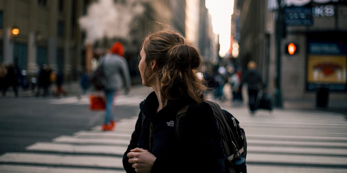 Eine junge Person steht im Vordergrund des Bildes, den Blick abgewendet Richtung Stadt. Im Hintergrund sind in der Unschärfe eine Kreuzung mit weiteren Menschen und Leuchtreklame zu erkennen.