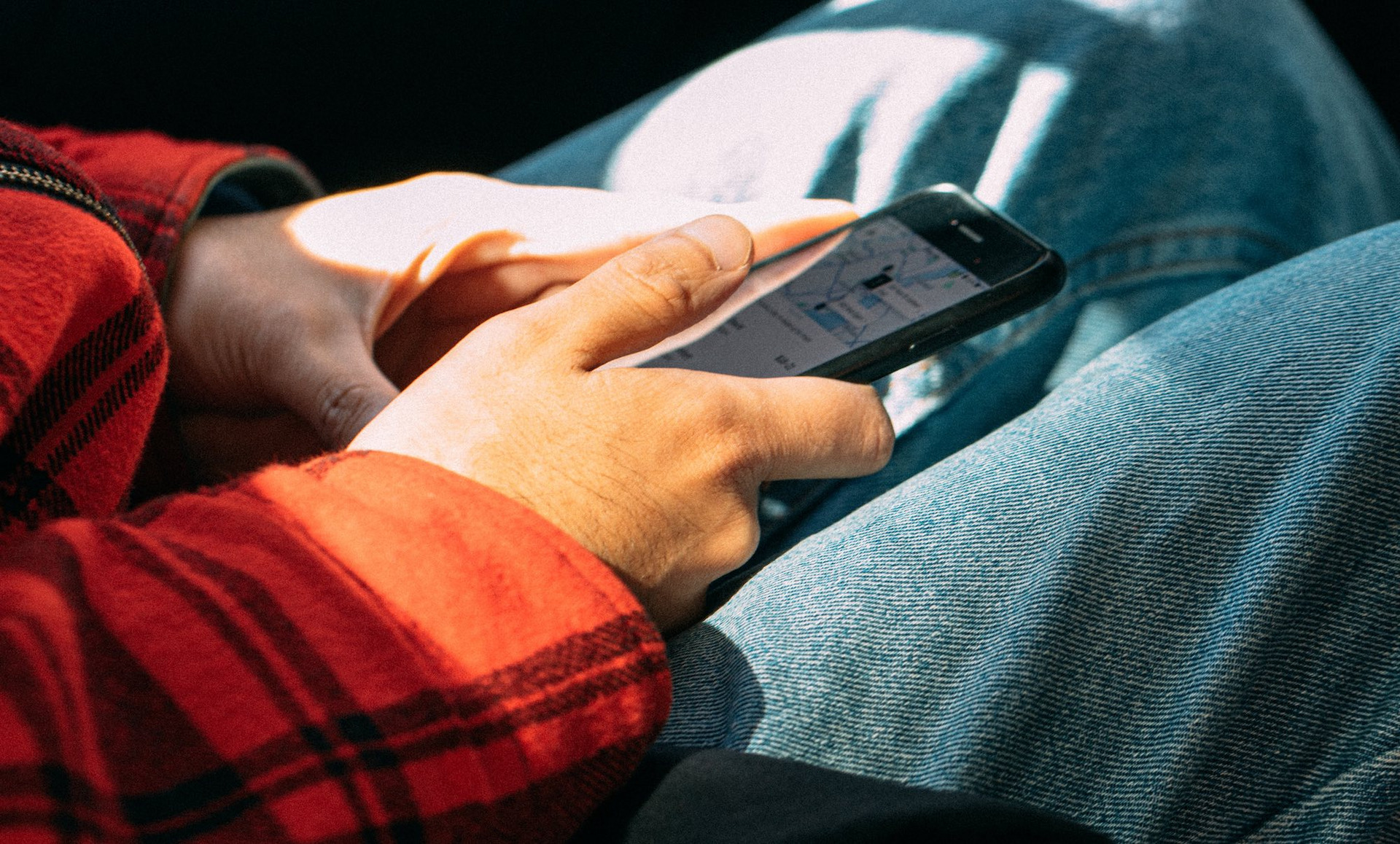 Warmes Sonnenlicht fällt durch die Scheibe eines Autos auf den Passagier. Wir sehen lediglich seine Hände, die ein Smartphone halten. Der Daumen ist zum Klick gehoben, auf dem Display ist eine Karte abgebildet.