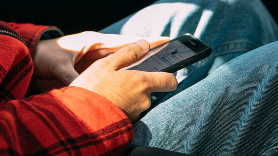 Warmes Sonnenlicht fällt durch die Scheibe eines Autos auf den Passagier. Wir sehen lediglich seine Hände, die ein Smartphone halten. Der Daumen ist zum Klick gehoben, auf dem Display ist eine Karte abgebildet.