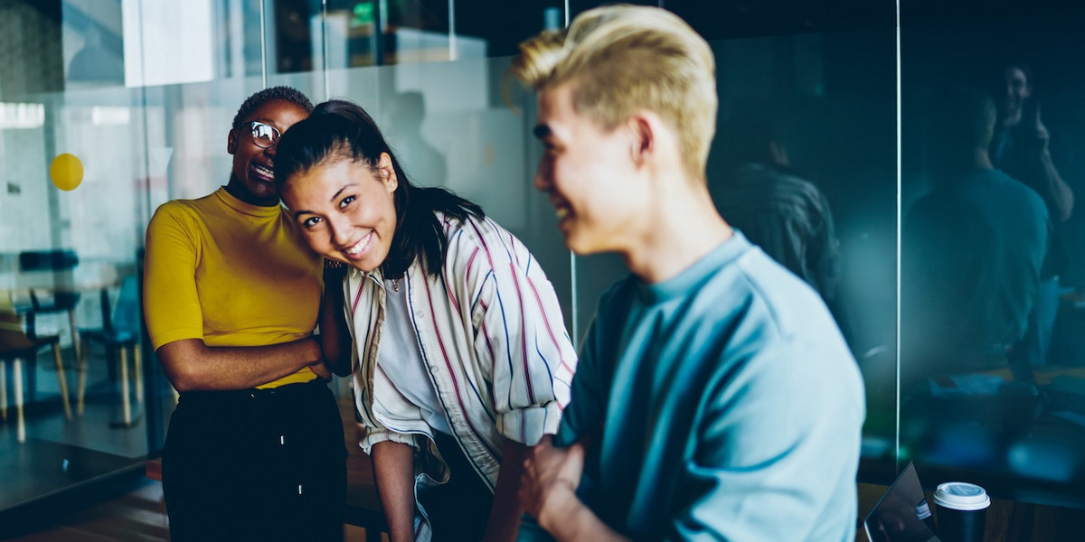 Drei junge Menschen stehen in einem modernen Büro und lachen zusammen. Die Person links trägt ein gelbes Oberteil und eine Brille, die Person in der Mitte hat dunkles Haar, das zu einem Pferdeschwanz gebunden ist, und trägt eine gestreifte Bluse. Die Person rechts hat kurzes blondes Haar und trägt ein hellblaues Oberteil. Im Hintergrund sind Glaswände und eine reflektierende Oberfläche zu sehen. Auf einem Tisch steht ein Kaffeebecher. Die Szene wirkt lebendig und fröhlich.