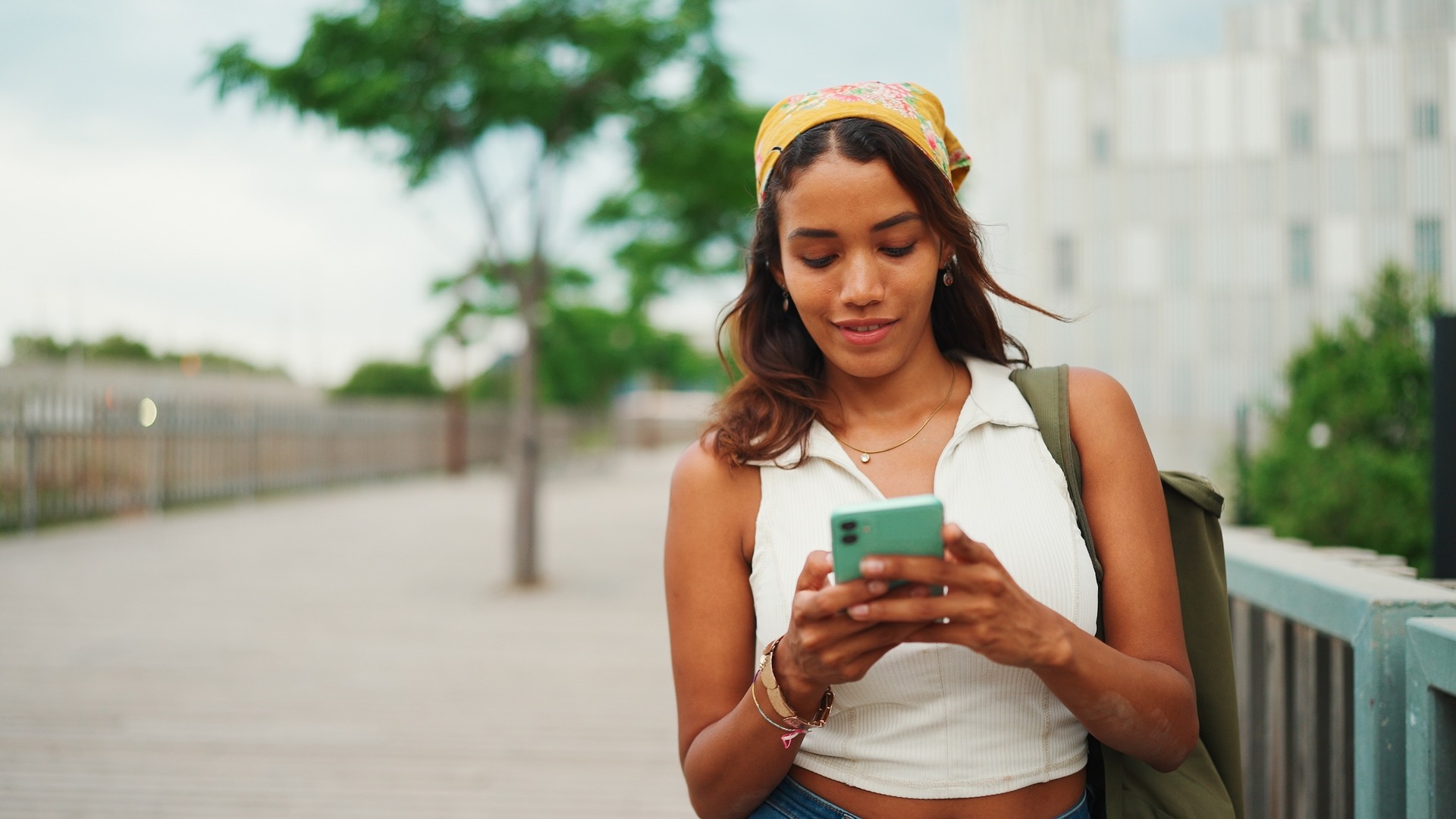 Eine junge Frau steht draußen auf einem Gehweg und schaut auf ihr Smartphone. Sie trägt ein ärmelloses Top und eine Tasche über der Schulter, im Hintergrund sind Bäume und Gebäude zu sehen.