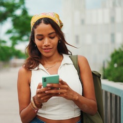 Eine junge Frau steht draußen auf einem Gehweg und schaut auf ihr Smartphone. Sie trägt ein ärmelloses Top und eine Tasche über der Schulter, im Hintergrund sind Bäume und Gebäude zu sehen.