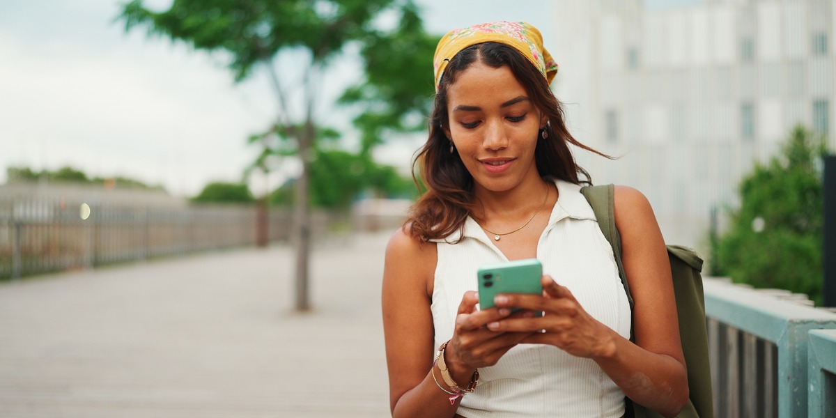 Eine junge Frau steht draußen auf einem Gehweg und schaut auf ihr Smartphone. Sie trägt ein ärmelloses Top und eine Tasche über der Schulter, im Hintergrund sind Bäume und Gebäude zu sehen.