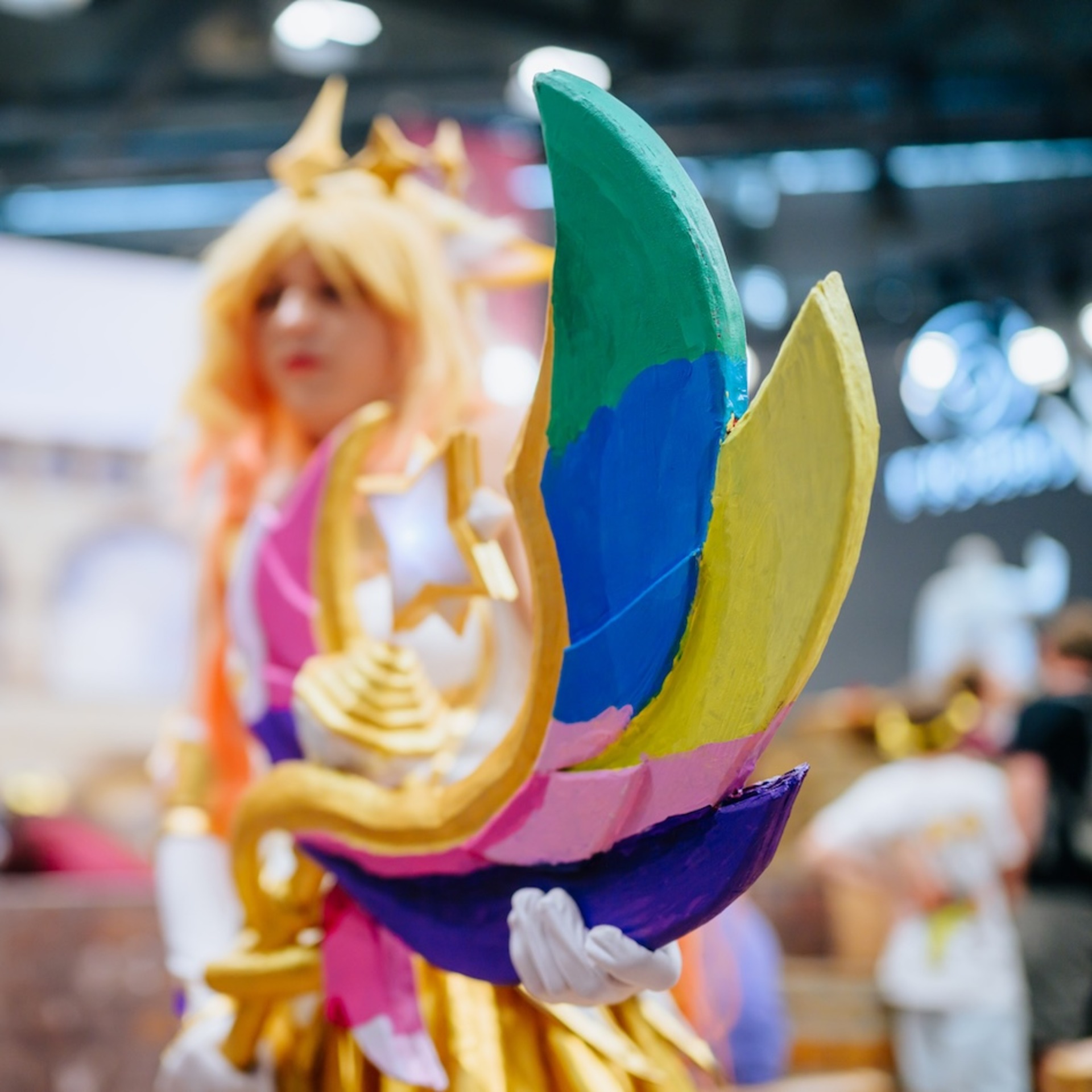 Eine goldene Krone mit bunten Federn in Grün, Blau, Gelb, Rosa und Lila steht im Vordergrund, dahinter sind unscharf Cosplay-Besucherinnen in Kostümen zu sehen. Die Szene wurde auf einer Convention oder Messe aufgenommen.