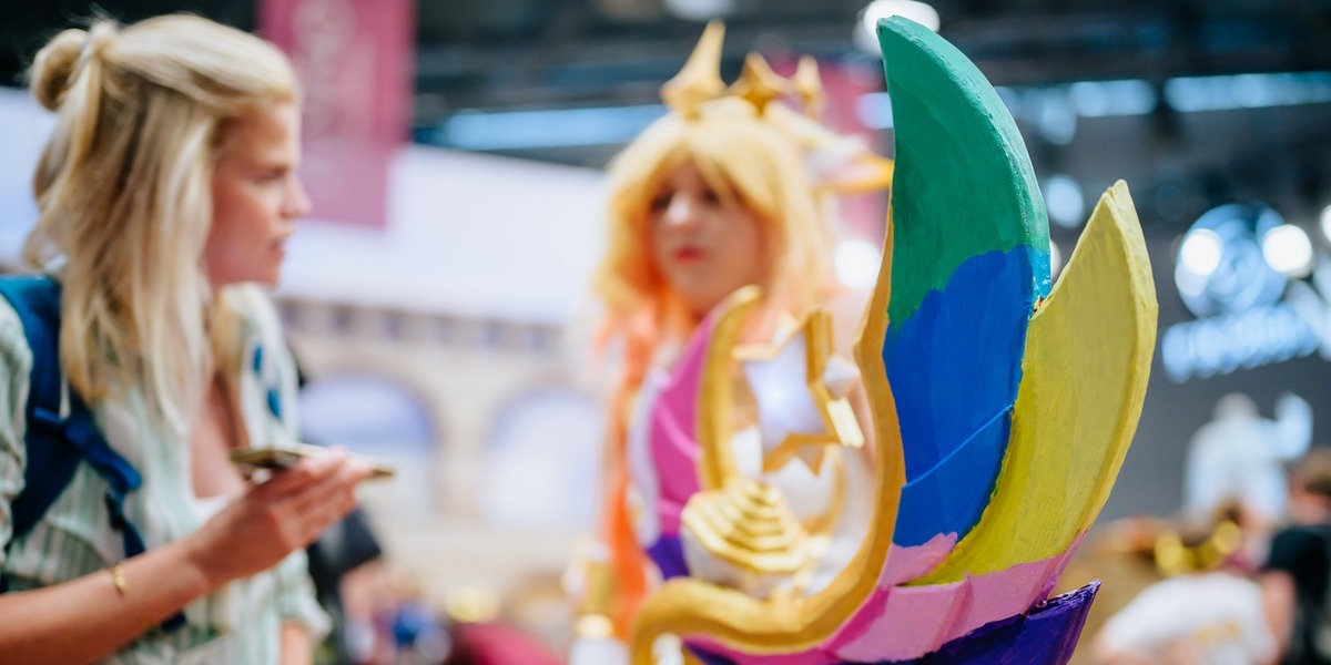 Eine goldene Krone mit bunten Federn in Grün, Blau, Gelb, Rosa und Lila steht im Vordergrund, dahinter sind unscharf Cosplay-Besucherinnen in Kostümen zu sehen. Die Szene wurde auf einer Convention oder Messe aufgenommen.