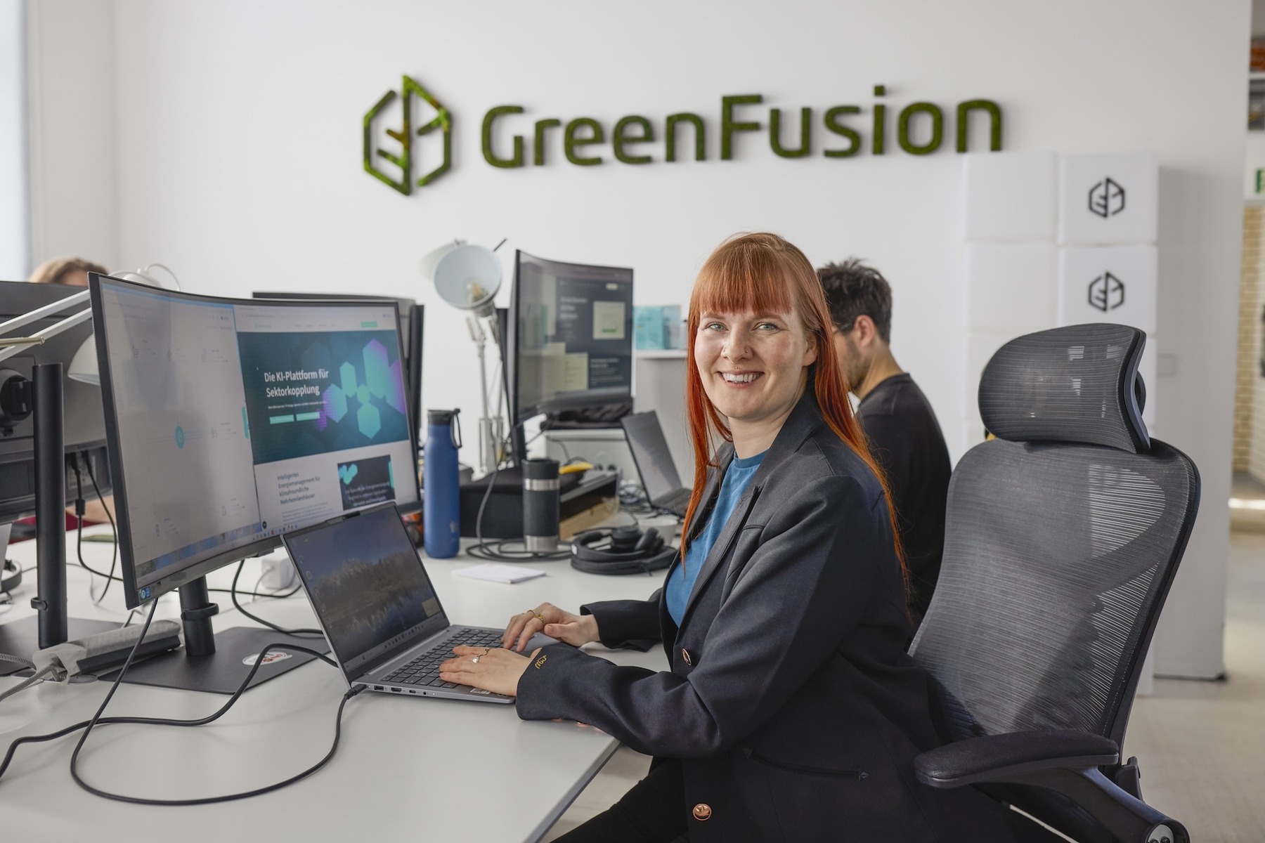 Eine Frau sitzt in einem Büro an einem Schreibtisch und lächelt in Richtung Kamera. Im Hintergrund ist eine weitere Person an einem Schreibtisch zu sehen. An der Wand ist die Aufschrift „GreenFusion“ zu sehen.