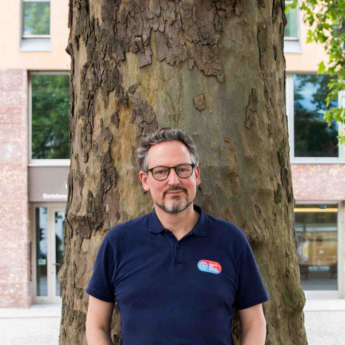 Eckart von Hirschhausen, ein Mann mittleren Alters, steht vor einem großen Baum. Er trägt eine Brille, ein dunkelblaues Polohemd mit einem kleinen Logo auf der linken Brust und eine karierte Hose. Seine Hände sind in den Hosentaschen. Im Hintergrund ist das Haus Berlin der Bertelsmann Stiftung, ein modernes Gebäude mit großen Fenstern und einer Fassade aus roten Ziegelsteinen, zu sehen. Der Mann wirkt entspannt und freundlich.