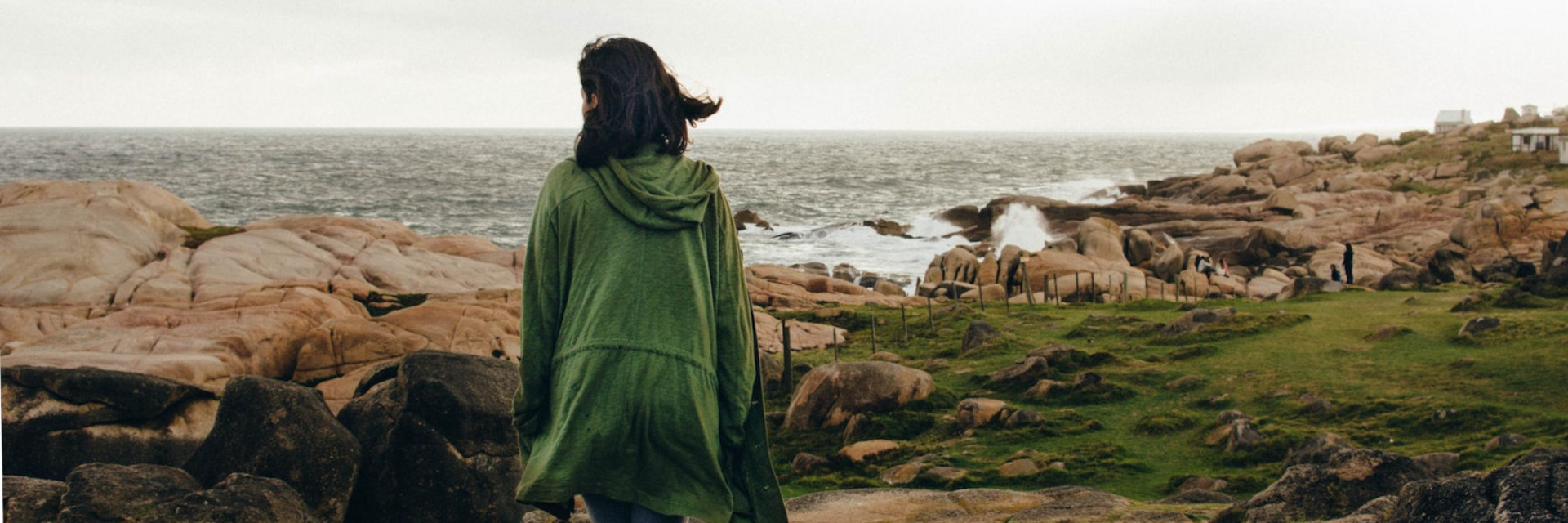 Eine Person in einem grünen Mantel läuft, uns den Rücken zugekehrt, den felsigen Strand von Cabo Polonio in Uruguay entlang, auf den tosende Wellen treffen.