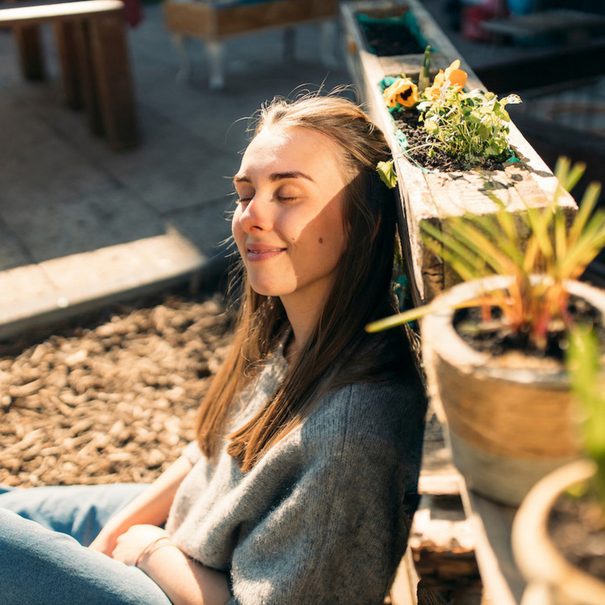 Eine junge Frau sitzt im Sonnenschein und lehnt sich entspannt an ein Hochbeet aus Holz, in dem Blumen und Pflanzen wachsen. Ihre Augen sind geschlossen, und sie genießt sichtlich die Wärme der Sonne auf ihrem Gesicht. Sie trägt ein graues Oberteil und blaue Jeans. Im Vordergrund befinden sich einige Topfpflanzen. Der Hintergrund zeigt eine gepflasterte Fläche und unscharfe Elemente eines Gartens oder einer Terrasse. Die Szene strahlt Ruhe und Zufriedenheit aus.