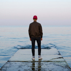 Ein Mann steht in sanftem rosa Abendlicht auf einem wellenumspielten  Steeg und blickt auf das Meer hinaus.