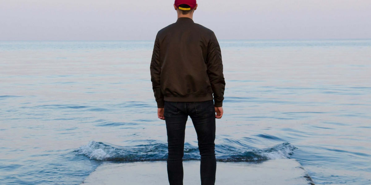Ein Mann steht in sanftem rosa Abendlicht auf einem wellenumspielten  Steeg und blickt auf das Meer hinaus.
