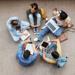 Luftaufnahme von sechs jungen Menschen, die im Kreis auf einem Betonboden sitzen und gemeinsam arbeiten. Sie haben Laptops, Tablets, Notizbücher und Taschen um sich verteilt.