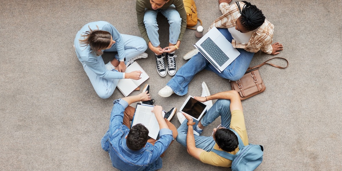 Luftaufnahme von sechs jungen Menschen, die im Kreis auf einem Betonboden sitzen und gemeinsam arbeiten. Sie haben Laptops, Tablets, Notizbücher und Taschen um sich verteilt.