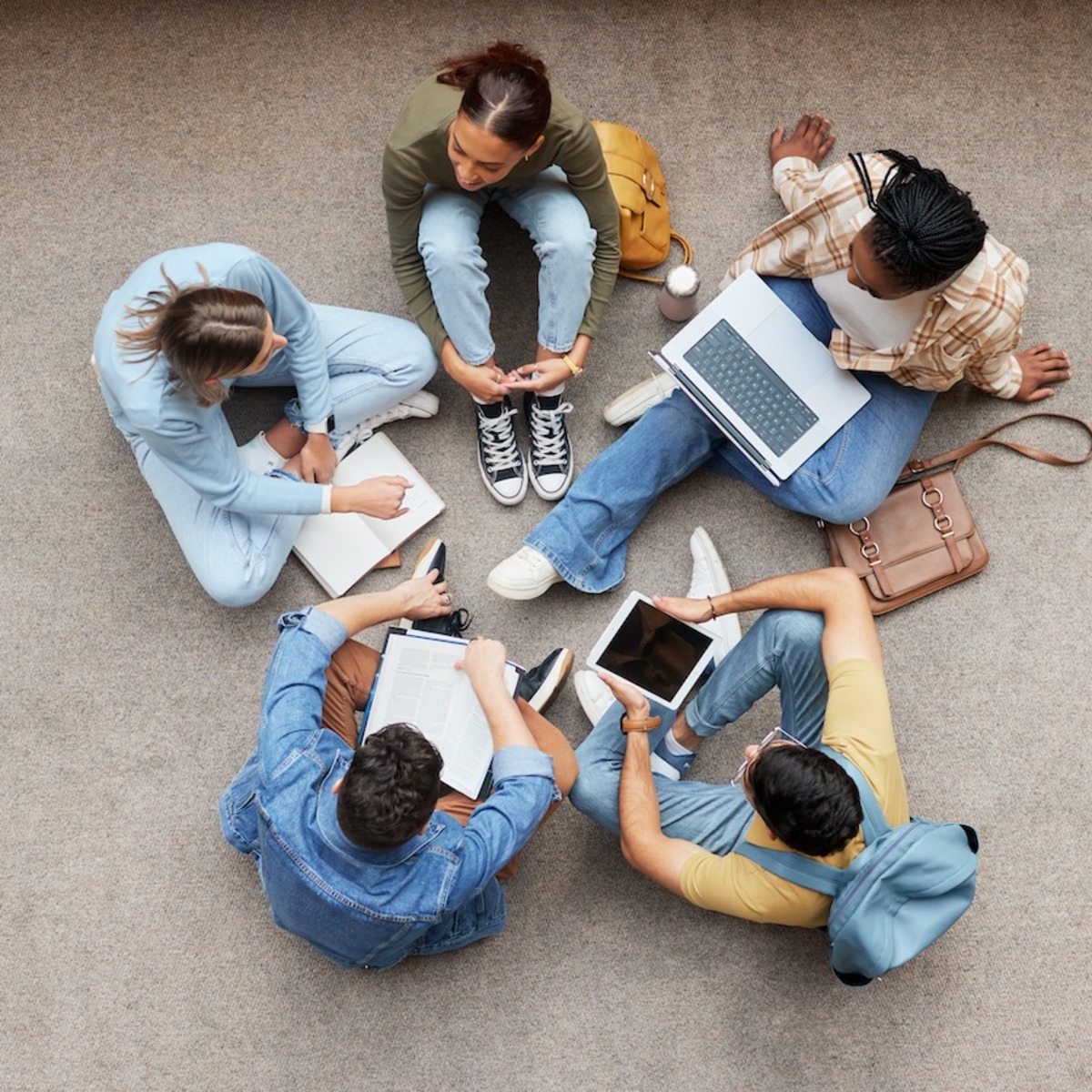 Luftaufnahme von sechs jungen Menschen, die im Kreis auf einem Betonboden sitzen und gemeinsam arbeiten. Sie haben Laptops, Tablets, Notizbücher und Taschen um sich verteilt.