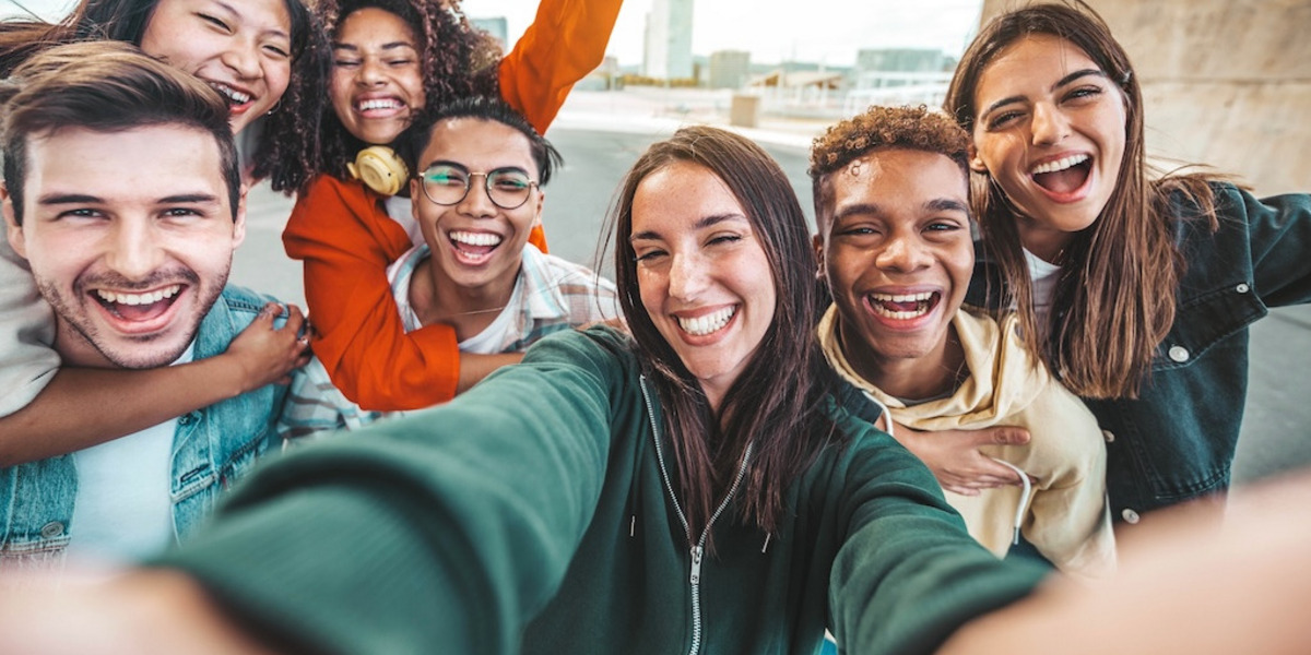 Eine Gruppe von sieben fröhlichen jungen Menschen macht ein Selfie, im Hintergrund ist die Kulisse einer Stadt zu sehen