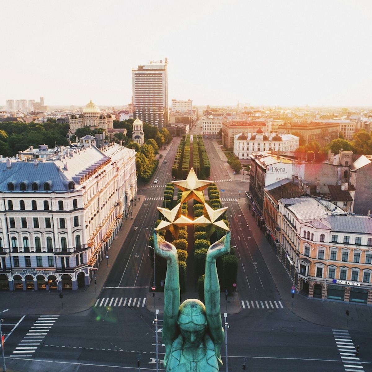 Luftaufnahme des Freiheitsplatzes in Riga mit breiter, baumgesäumter Allee und zentralem Monument mit goldenen Sternen, umgeben von historischen Gebäuden mit klassischer Architektur. Im Hintergrund ragt ein moderner Hochhausturm über die traditionelle Stadtsilhouette hinaus.