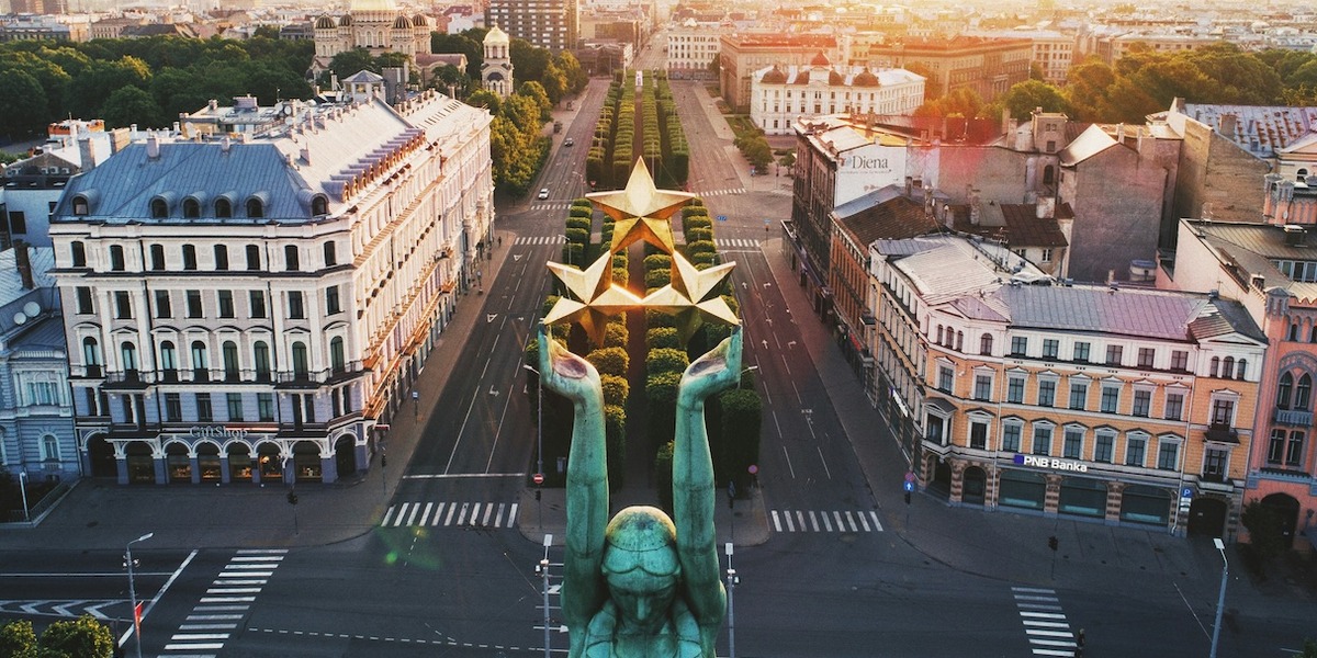 Luftaufnahme des Freiheitsplatzes in Riga mit breiter, baumgesäumter Allee und zentralem Monument mit goldenen Sternen, umgeben von historischen Gebäuden mit klassischer Architektur. Im Hintergrund ragt ein moderner Hochhausturm über die traditionelle Stadtsilhouette hinaus.