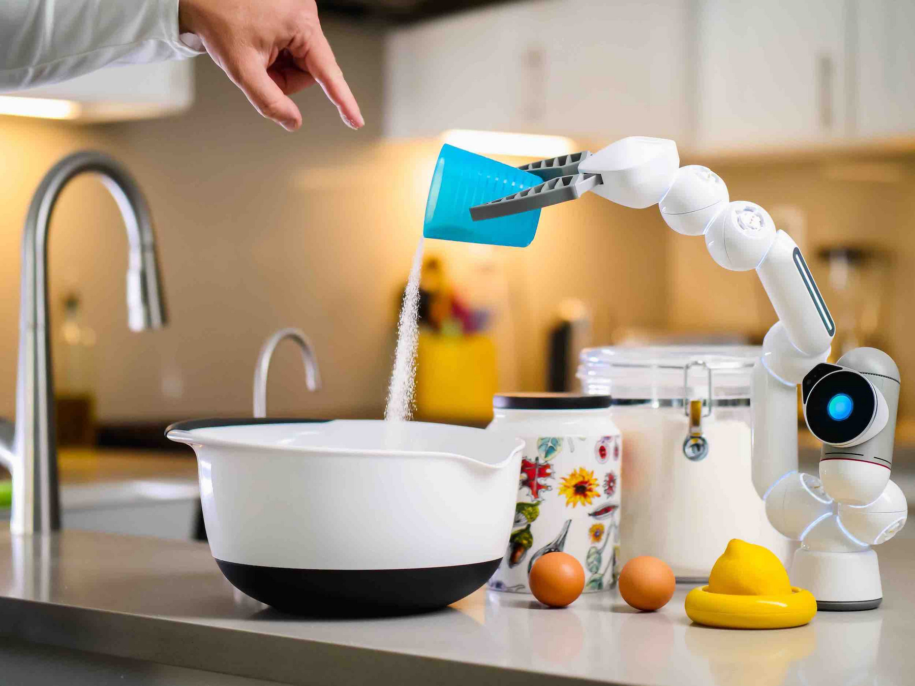 Ein Roboterarm steht auf einer Kochzeile und schüttet Zucker in eine Rührschüssel. Daneben ein Gefäß mit Zucker, zwei braune Hühnereier und eine halbe Zitrone. Am oberen Rand des Bildes ist eine Hand zu sehen, die mit dem Finger in die Rührschüssel zeigt.