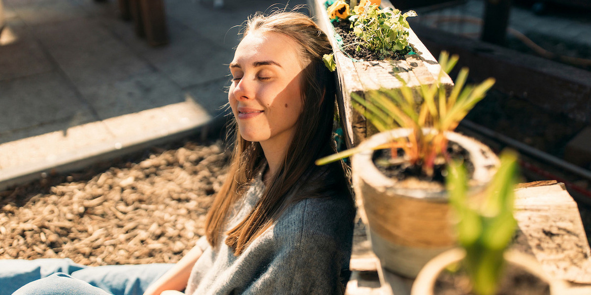 Eine junge Frau sitzt im Sonnenschein und lehnt sich entspannt an ein Hochbeet aus Holz, in dem Blumen und Pflanzen wachsen. Ihre Augen sind geschlossen, und sie genießt sichtlich die Wärme der Sonne auf ihrem Gesicht. Sie trägt ein graues Oberteil und blaue Jeans. Im Vordergrund befinden sich einige Topfpflanzen. Der Hintergrund zeigt eine gepflasterte Fläche und unscharfe Elemente eines Gartens oder einer Terrasse. Die Szene strahlt Ruhe und Zufriedenheit aus.