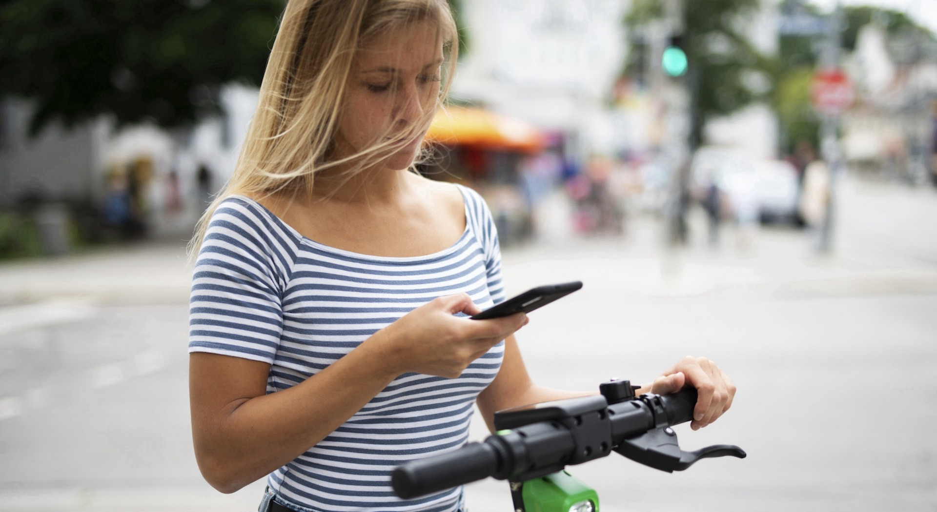 Das Foto zeigt eine junge Frau bei Tageslicht auf der Straße, der Hintergrund ist unscharf, aber man erahnt eine Kreuzung mit einer grünen Ampel in der Ferne. Die Frau hält ein Smartphone in der rechten Hand, ihre linke Hand umgreift den Lenker eines E-Rollers, auf dem sie steht.