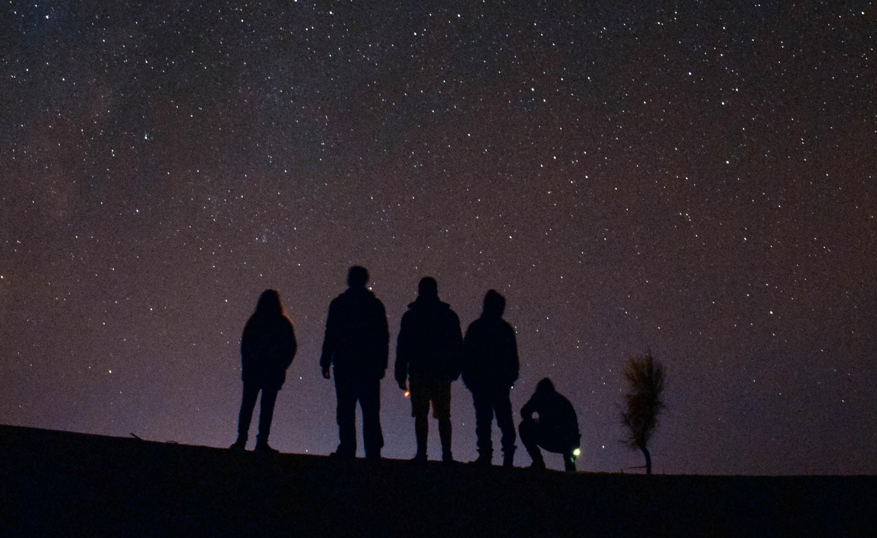 Sechs Menschen stehen auf einem Hügel vor sternenklarem Nachthimmel. Es ist tiefdunkel, man erkennt nur die Himmelskörper und die Silhouetten der Personen.