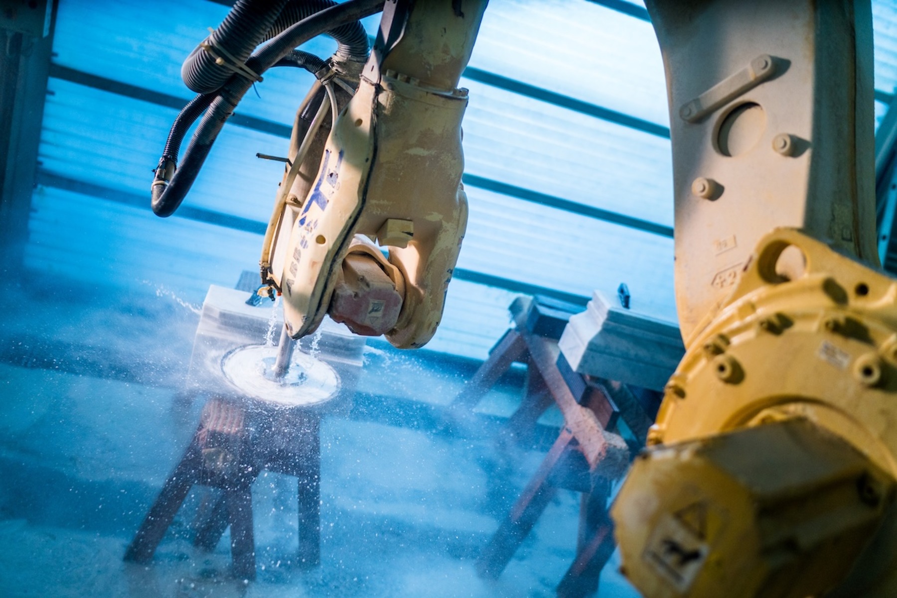 Ein 5-Achs-Roboter bearbeitet präzise eine Marmorplatte, wobei Staub und Späne aufgewirbelt werden. Im Hintergrund sind weitere Roboterkomponenten in blauer industrieller Beleuchtung zu sehen.