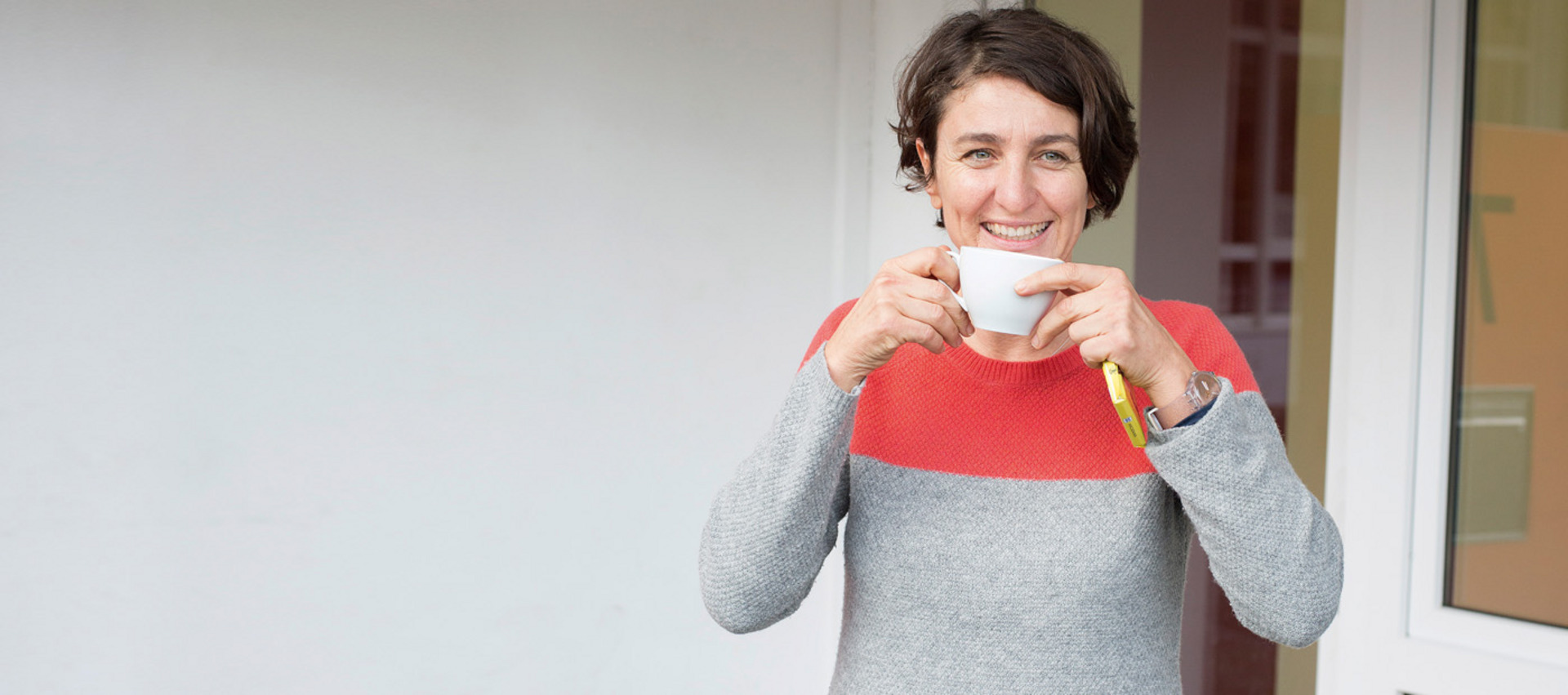 Christiane Falge trinkt aus einer Kaffeetasse und lacht in die Kamera.