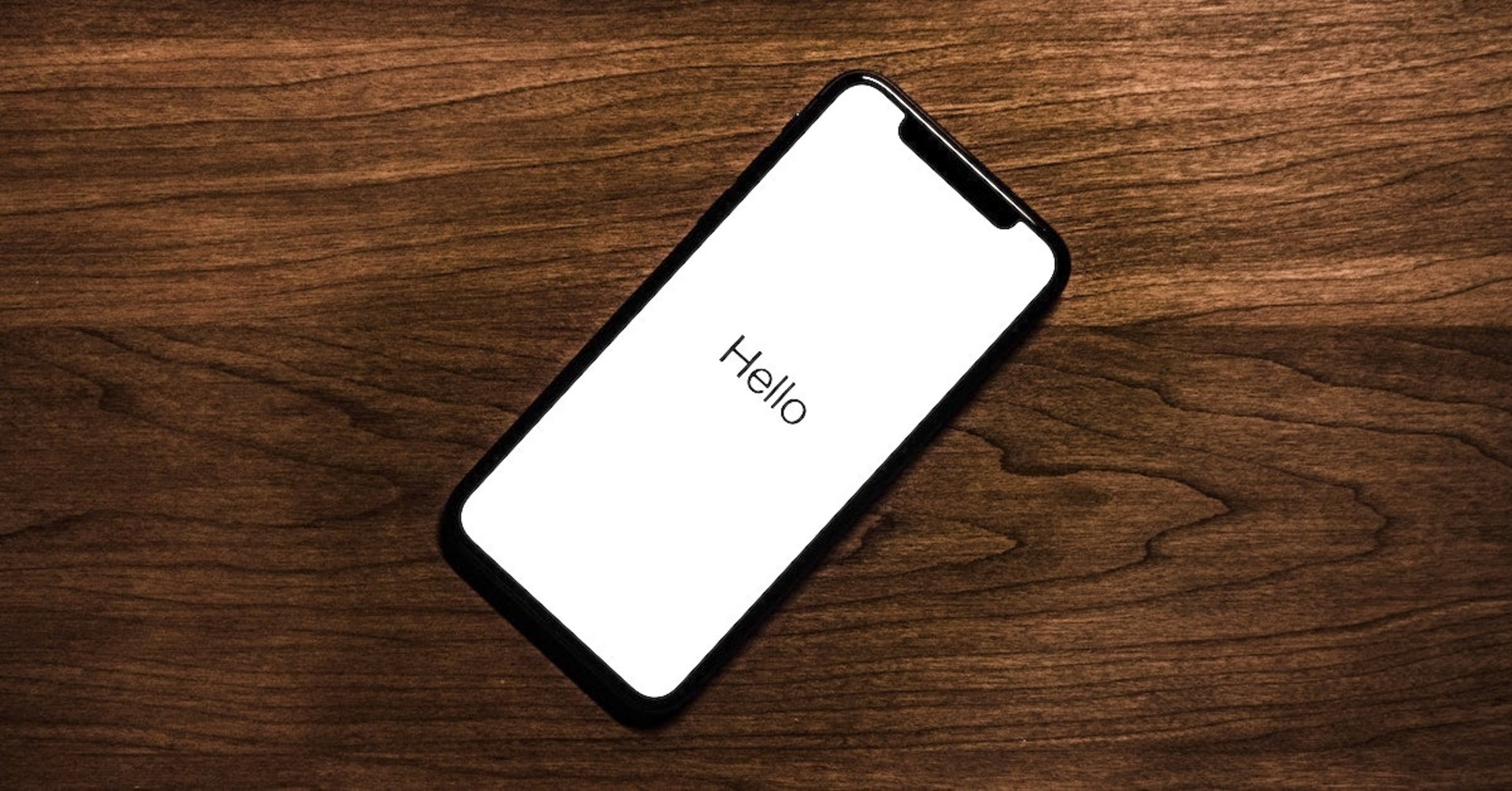 Ein Smartphone mit dem Text "Hello" auf dem Schirm liegt auf einem Holztisch.