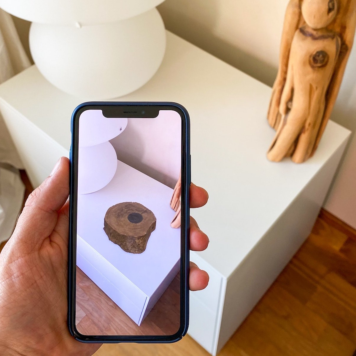 Das Foto zeigt eine Hand, die ein Smartphone vor eine Kommode hält. Auf der Oberfläche erscheint mittels Augmented Reality ein Designobjekt in Form einer Holzskulptur.