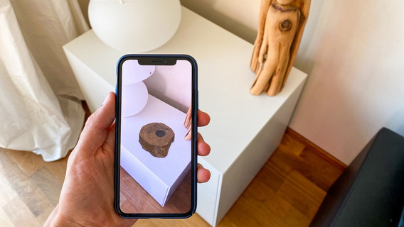 Das Foto zeigt eine Hand, die ein Smartphone vor eine Kommode hält. Auf der Oberfläche erscheint mittels Augmented Reality ein Designobjekt in Form einer Holzskulptur.