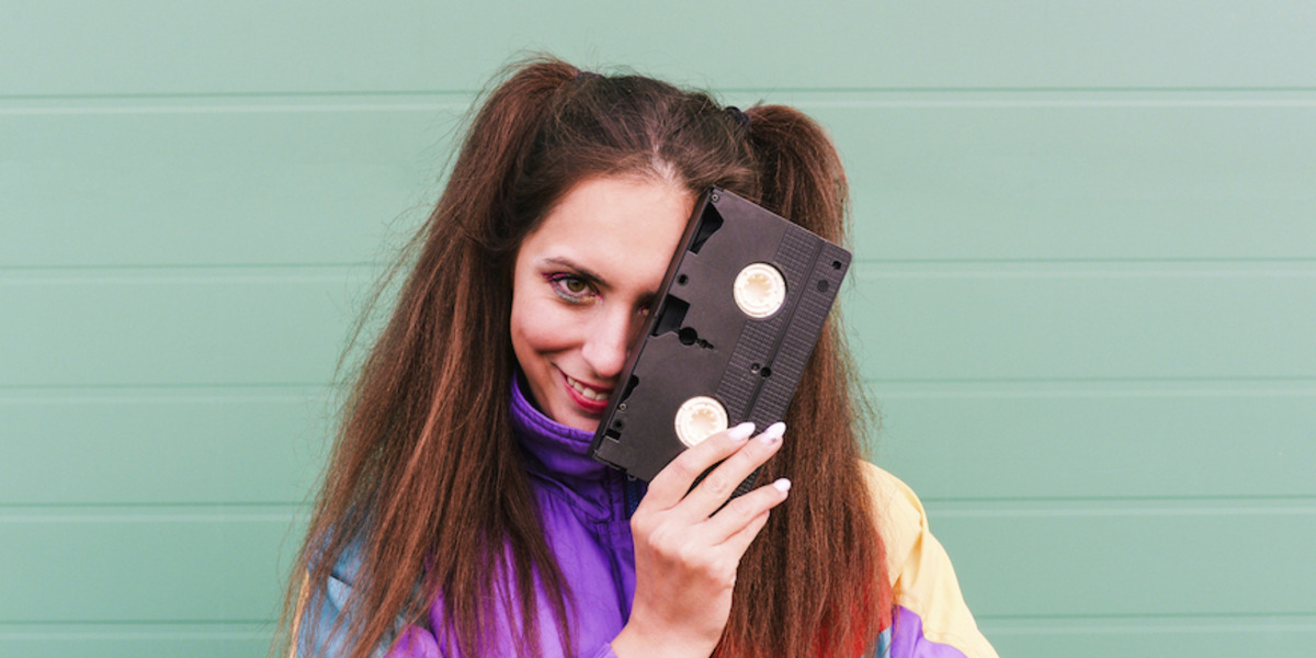 Eine junge Frau in einer bunten Sportjacke hält sich eine VHS-Kassette vor das Gesicht