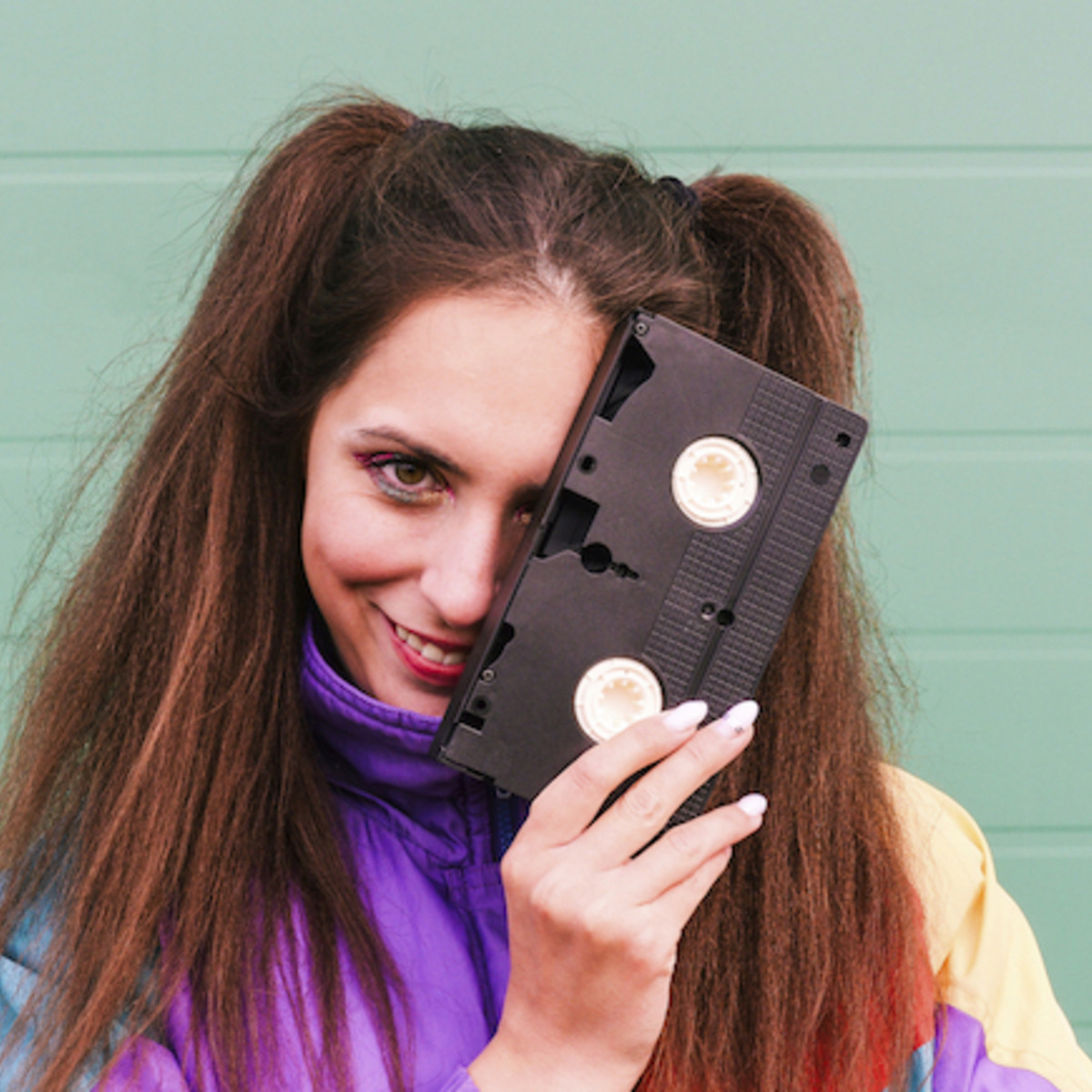 Eine junge Frau in einer bunten Sportjacke hält sich eine VHS-Kassette vor das Gesicht