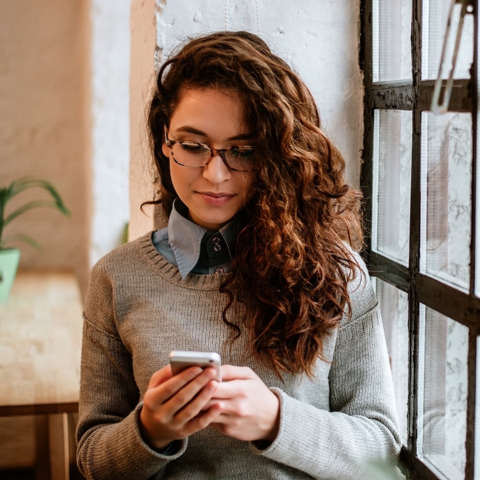 Eine junge Frau mit lockigem Haar schaut intensiv auf ihr Smartphone, das sie mit beiden Händen hält. Im Hintergrund ist ein Wohnraum zu sehen.
